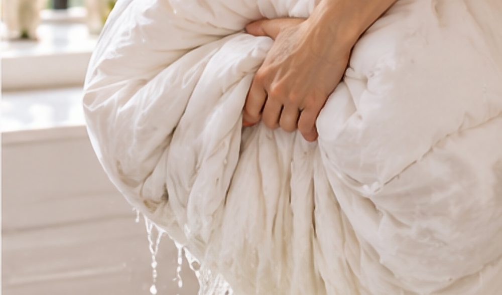 Hands lifting a soaked white duvet out of a bathtub as water streams off it