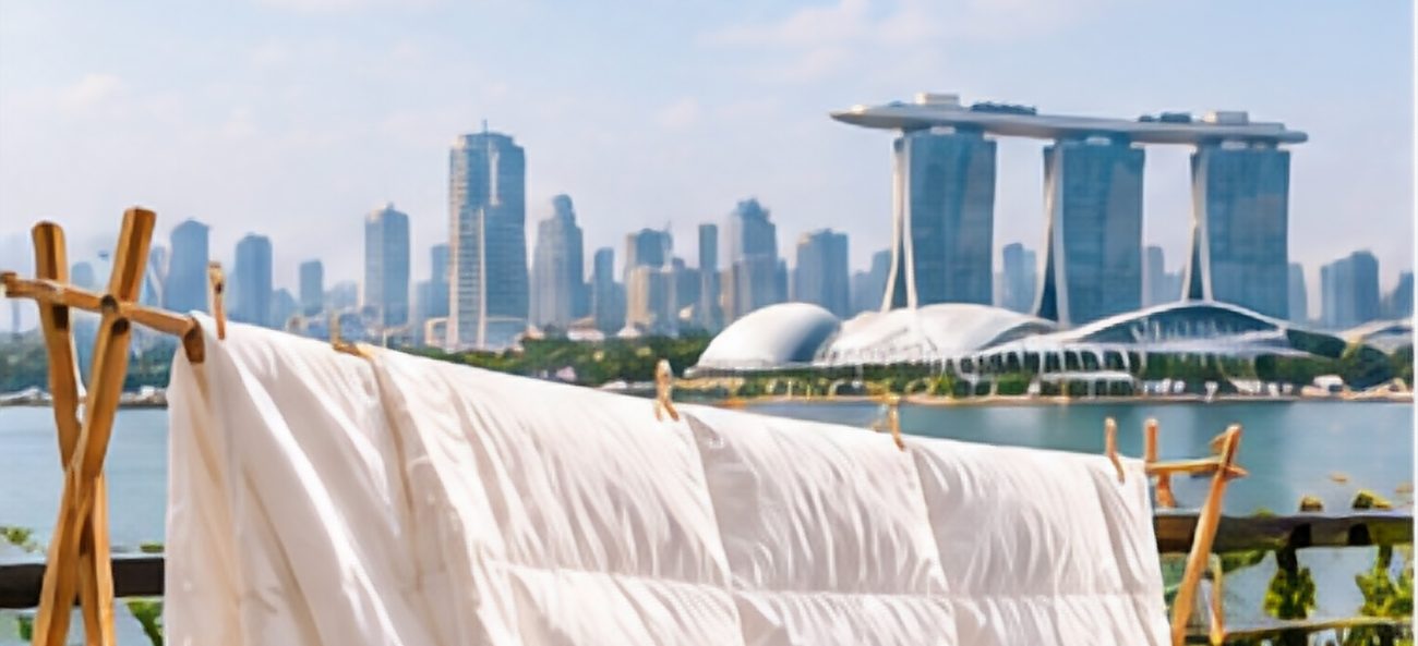 A white quilted duvet draped over a wooden drying rack on a sunny terrace with the Marina Bay skyline behind